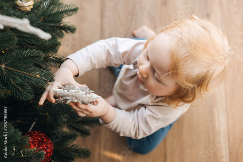 Happy little toddler decorating a Christmas tree at home, viewed from above. Cute child reaching for festive ornaments during Christmas preparation, enjoying a cozy family holiday tradition.