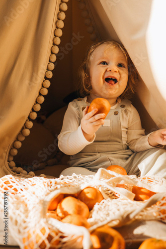 Cute toddler sitting inside a cozy blanket fort and exploring fresh oranges from a reusable mesh shopping bag at home. Candid childhood lifestyle scene with healthy food and imaginative play.