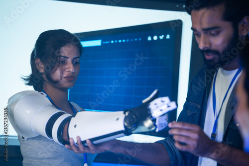 A diverse team tests a robotic arm prototype in a modern tech workshop. Engineers collaborate closely while examining the mechanical limb design against a large digital screen in the background.