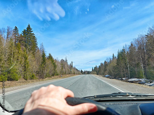 Fotografie Driver hand and steering wheel frame an open forest road in spring, with vivid b