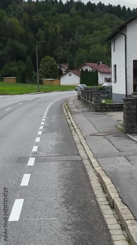 Cycling through a town street, camera view from the cyclist’s perspective, urban road and buildings visible. A road is wet from rain. There are houses on the side and green fields in the background. T