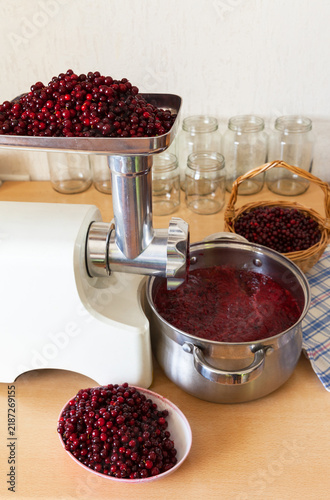 Making lingonberry jam. Grinding ripe red berries in an electric meat grinder; a wicker basket of berries and jars on the kitchen table. Preparing winter supplies