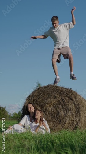 A young mother and her 7 year old daughter, wearing white dresses are sitting on a green meadow next to a stack of hay on a sunny day. A 14 year old boy is jumping on the stack of hay and laughing.