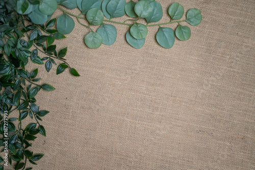 Rustic flat lay background with a corner frame of green eucalyptus and ivy leaves on natural burlap fabric texture and ample copy space