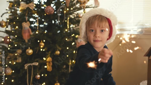 Happy boy in santa hat playing with sparkler near christmas tree