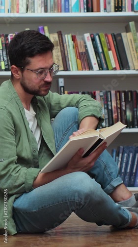 Caucasian student man reading book, sitting among stack of books on library floor, side view.