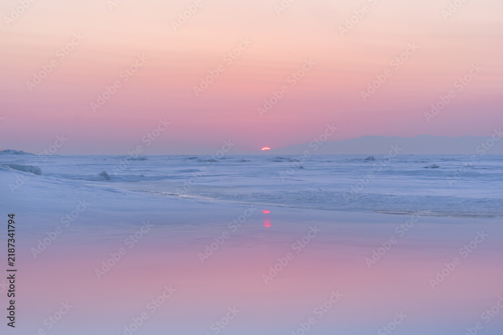 Obraz premium Pastel sunset over a frozen sea with snow-covered ice and distant hills, creating a calm and peaceful winter landscape.