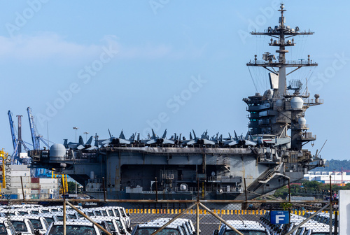 Fighter jets line the aircraft carrier. Large cranes tower over the aircraft carrier. A military aircraft carrier rests at the harbor.