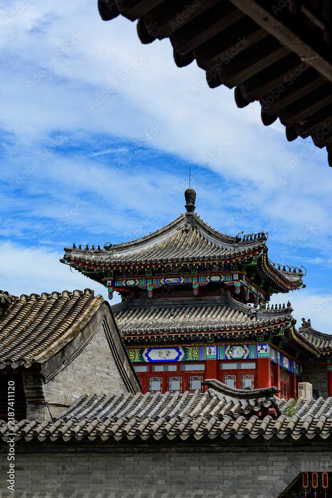 Fototapeta premium Framed view of an ornate multi-tiered ancient Chinese pavilion, vibrant painted dougong brackets, grey tiled roofs, bright blue sky with wispy clouds.