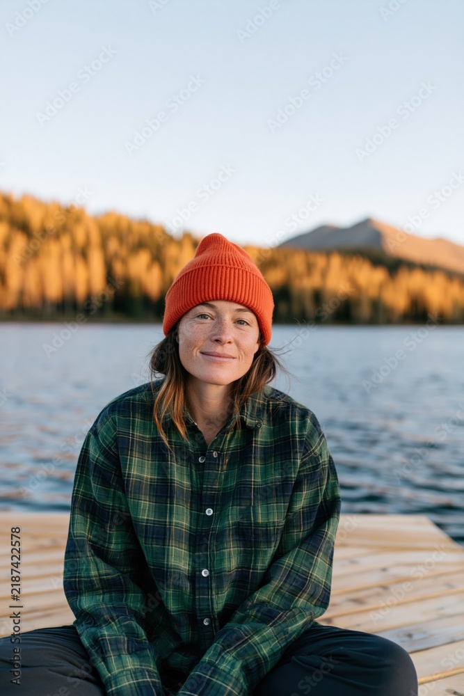 Fototapeta premium Young woman in orange beanie and plaid shirt on lakeside dock with autumn trees for outdoor portrait background