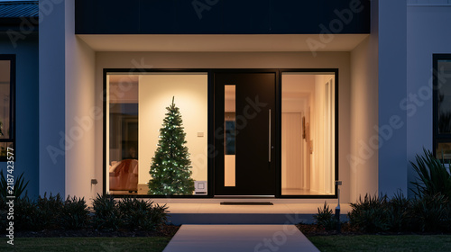 Modern house exterior at night with illuminated Christmas tree visible through the entrance, luxury property and holiday home. 
