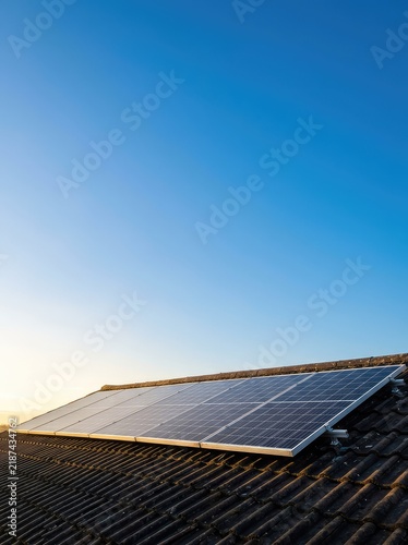 A row of solar panels across a rooftop, capturing the warm glow of early morning sunlight with a clear, rich blue sky in the background, Rooftop, Sunrise, Roof
