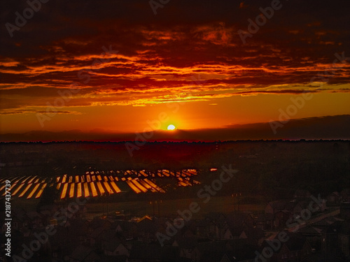 Aerial view of a solar farm at sunset with glowing panels and a residential area in the foreground under a dramatic orange sky in Lisburn, Northern Ireland, United Kingdom.
