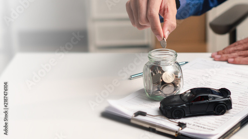 Obraz na plátně Person saving coins in a glass jar beside a model car and finance documents, symbolizing auto loan planning, vehicle purchase, budgeting, ownership costs, investment, and financial security