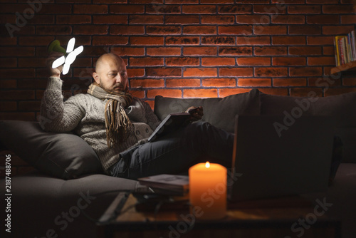 Ukrainian man on sofa in warm clothes reading book using portable led lamp and candle light during power outage in cold dark home caused by Russian attacks on energy grid