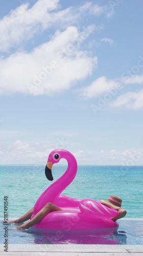 Funny travel vacation holidays concept. Woman in pool by beach sunbathing in bikini in inflatable pink flamingo toy float relaxing in luxury. Raw vertical video of real woman model without makeup