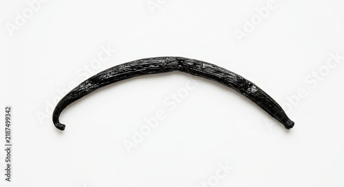 Single black dried vanilla bean pod on white surface.