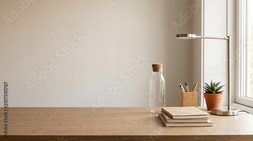Minimalist desk workspace featuring stacked neutral notebooks, small succulent plant, glass bottle, wooden pen holder, and modern silver lamp light