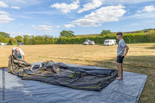 Two men set up a large tent on a campsite field, with caravans, green hedges and a bright blue sky with fluffy clouds in the background.