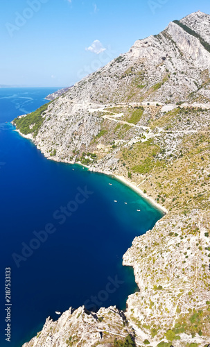 Aerial drone view of a scenic Adriatic coastline near Pisak, Croatia, featuring dramatic rocky mountains, a turquoise secluded bay, deep blue sea and small yachts in crystal clear water.