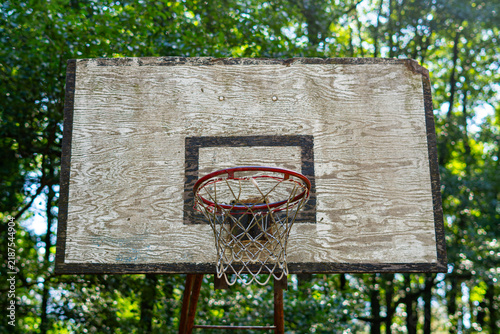 Bild auf Leinwand Old wooden basketball backboard with worn, peeling white paint, a red metal rim, and a net, set against a background of dense green tree foliage in summer light