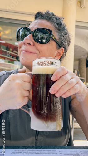 A woman wearing sunglasses holds a frosted mug of dark beer while sitting at an outdoor table.