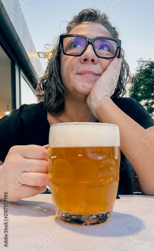 A woman wearing glasses looks away while holding a glass of beer outdoors.