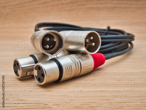 XLR Audio Cables on Wooden Background - Four shiny XLR connectors and a coiled black audio cable lie on a light wooden surface, ready for professional sound equipment connections.
