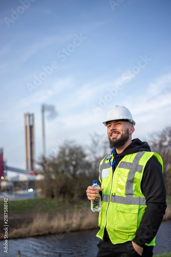 Industrial worker wearing hard hat and safety vest, holding water bottle at a factory site