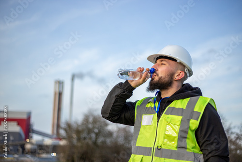Industrial worker hydrating with water bottle, wearing hard hat and safety vest at factory site