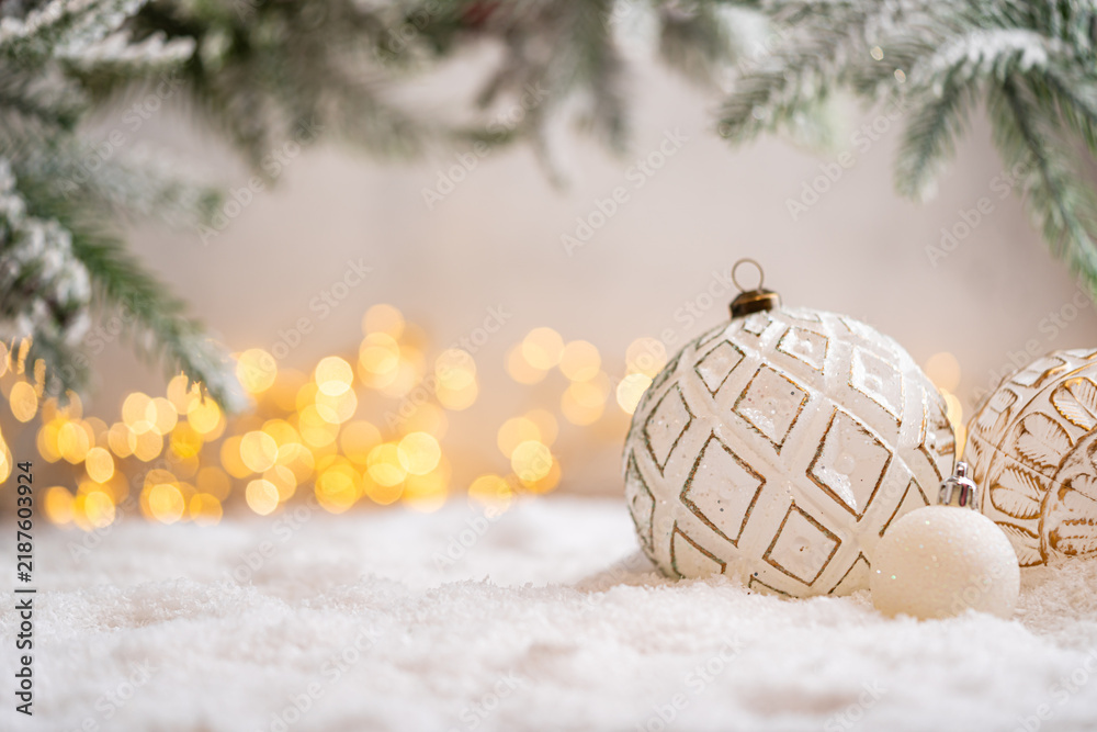Naklejka premium A Christmas background featuring an elegant white bauble etched with a golden diamond pattern beneath frosted pine on snow, softly lit by warm twinkling lights with generous copy space.