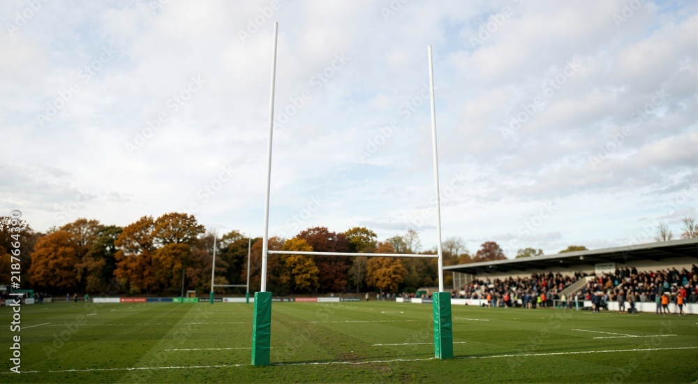Fototapeta premium Rugby goal posts on a green grass field. Stadium stand with spectators and autumn trees under cloudy sky