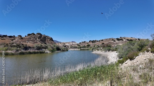 Fotografía A small lake nestled among dry hills reflects the clear sky, surrounded by sparse vegetation and rocky slopes, creating a peaceful contrast between water and arid terrain
