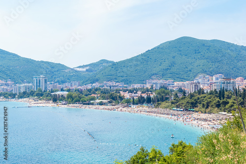 Scenic panoramic view of Slovenska Plaza beach bay in Budva, Montenegro. Sea coastline with green mountains and cityscape under bright blue sky