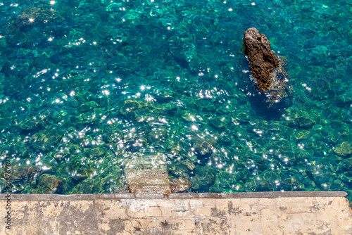 Sunlight glares on crystal clear turquoise sea water near a stone pier in Budva, Montenegro. Top view of transparent adriatic sea bottom