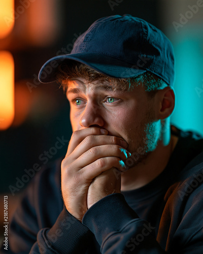 Anxious Young Man in Dim Light: A young man with curly hair, wearing a blue cap, clasps his hands over his mouth, his eyes wide with apprehension and distress.