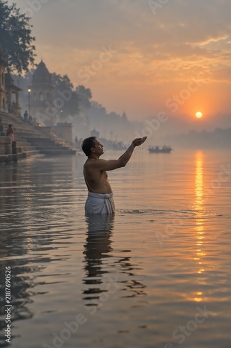Indian man performing sacred ritual bath offering water at sunrise on holy river ghat. Misty morning with temple silhouette and golden sun reflection.