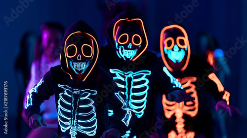Front-facing portrait of three people wearing black hooded Halloween costumes printed with glowing neon skeleton bones, fluorescent skull masks outlined in electric orange, cyan an