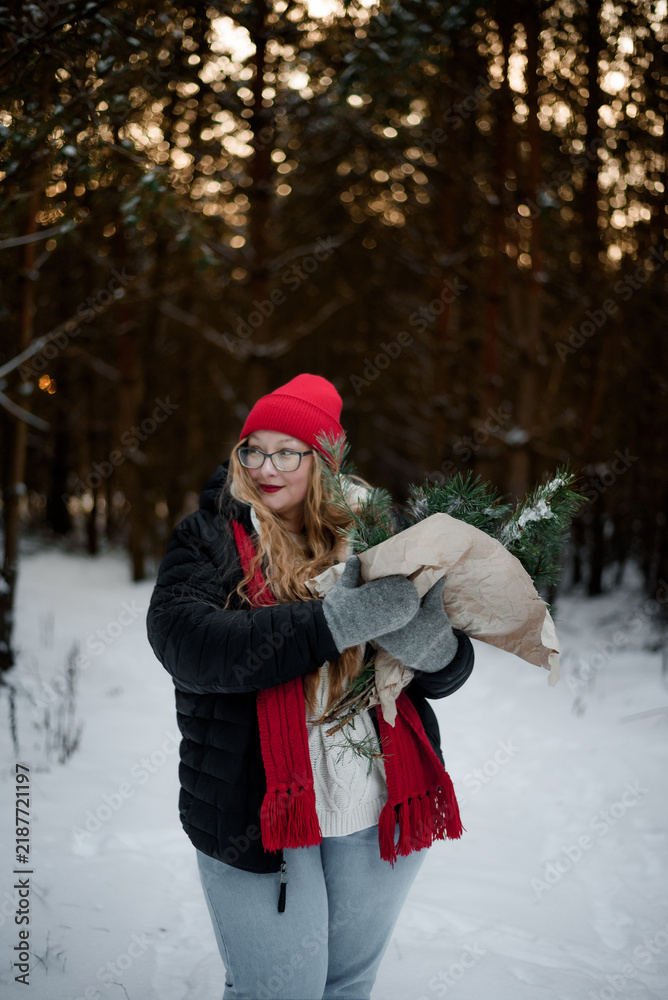 Fototapeta premium Smiling young woman in winter outfit holding pine branches outdoors in snowy forest. Concept of Christmas holidays, winter lifestyle, happiness, seasonal nature and festive mood.
