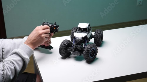Person's hands holding a black radio remote control, manipulating a white and black toy rock crawler car with large tires sitting on a white table, engaging in hobby and leisure activity indoors
