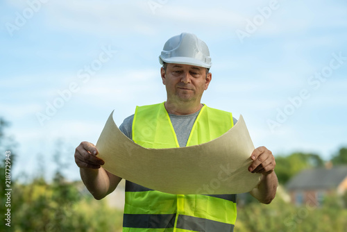 Fotografie A diligent worker, wearing a safety helmet and bright vest, carefully examines construction blueprints in a lush, green area
