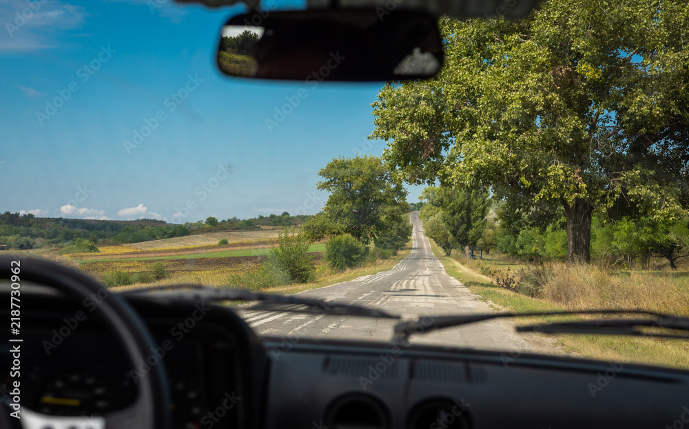 Obraz premium View through a car windshield of a rural country road in Romania, lined with trees and bordered by farmland, stretching toward distant hills under a clear blue sky.