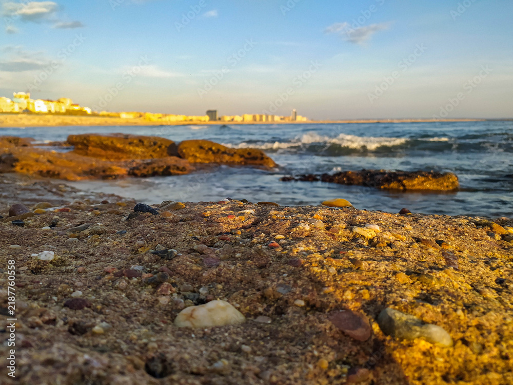 Fototapeta premium A breathtaking lowangle view of a rocky shore at golden hour. Warm sunlight illuminates the coast, while ocean waves meet a distant city skyline. Ideal for travel and lifestyle concepts.