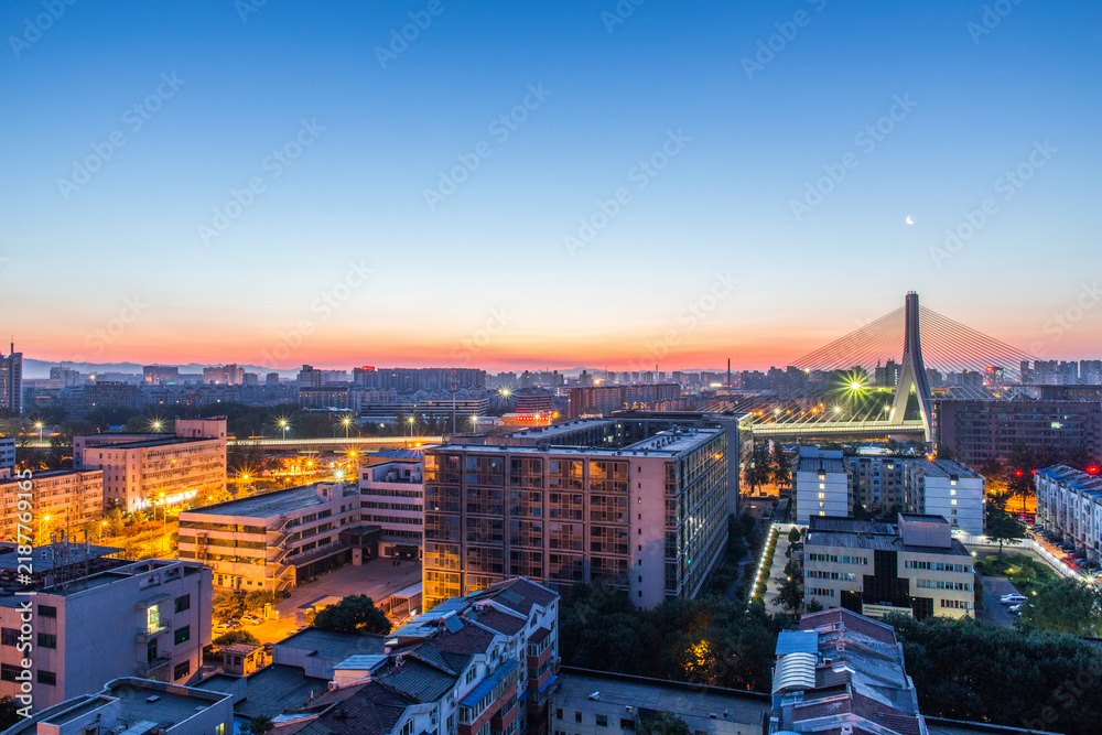Fototapeta premium A panoramic urban view at dawn, featuring a cable-stayed bridge, lit buildings, and a gradient sky with a visible moon.