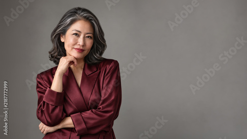 A mature woman with wavy gray hair poses in a burgundy satin jacket, resting her chin on one hand and smiling gently against a gray backdrop.