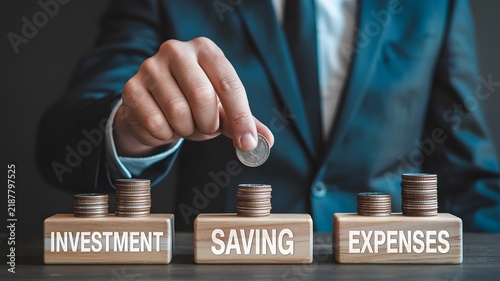 Financial management and wealth planning concept. Hand of a businessman placing a silver coin onto a stack atop wooden blocks labeled investment, saving, and expenses. Corporate finance strategy
