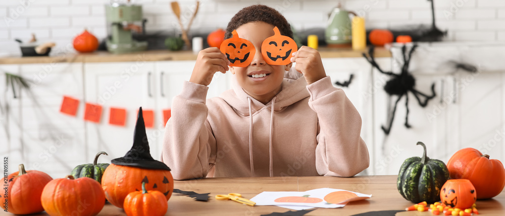 Fototapeta premium Halloween celebration and childhood joy, happy boy holding paper pumpkin masks over eyes while crafting at home kitchen table with autumn decorations