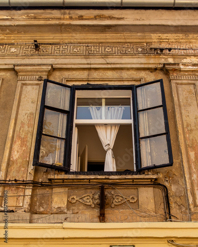 Foto An open window with white curtains on a weathered historic building facade, left open to allow fresh air and natural light into the interior of the vintage structure