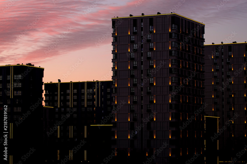 Fototapeta premium Night city background with illuminated lights on modern architecture — real estate concept. Beautiful atmospheric urban sunset sky with pink colorful clouds over dark residential block exterior.