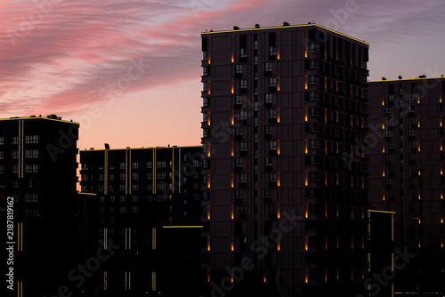Night city background with illuminated lights on modern architecture — real estate concept. Beautiful atmospheric urban sunset sky with pink colorful clouds over dark residential block exterior.